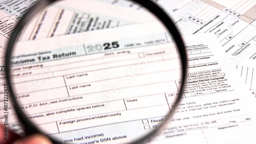 A person examines various tax documents and 1040 with with a magnifying glass. The focus is on details like numbers and sections relevant to tax filing. It takes place during tax season at home.