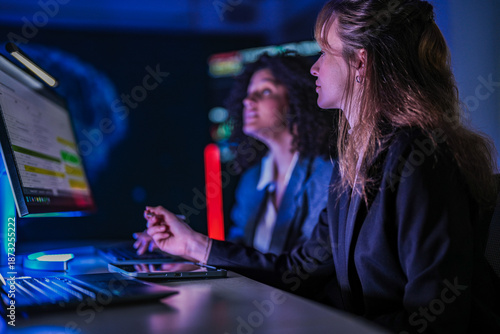 Dedicated business team working late on corporate project. Professional women analyzing digital performance data on screens in modern dark blue office at night.