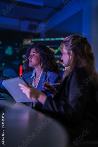 Dedicated business team working late on corporate project. Professional women analyzing digital performance data on screens in modern dark blue office at night.