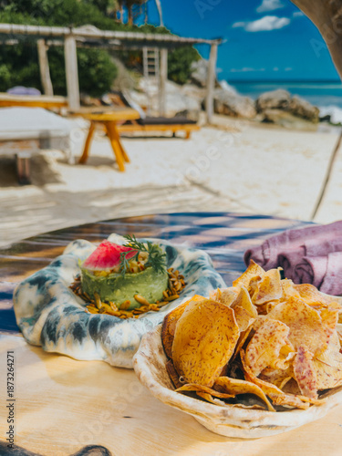 Obraz na płótnie Fresh guacamole served with crispy root vegetable chips on a wooden table overlooking a sandy beach in Tulum, Mexico