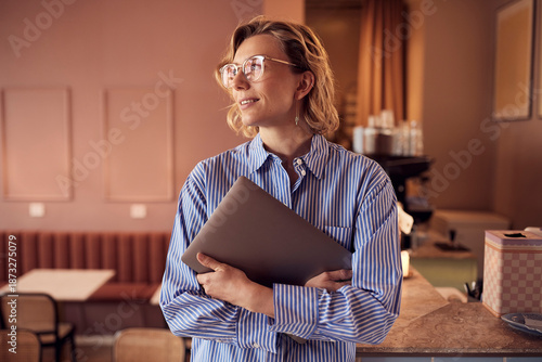 Casual cafe scene with a woman holding a laptop, looking confident and focused on work. small shop owner.