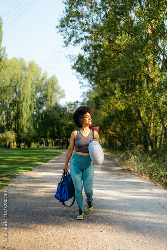 Afro woman walking on a park path, carrying her yoga mat and gym bag