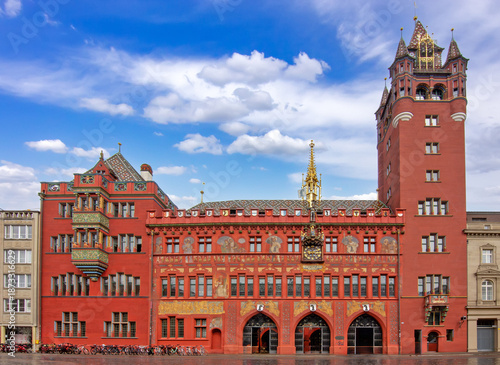 Historic red town hall in Basel with ornate facade, clock tower, painted murals, and bicycles in the city square under a blue sky.