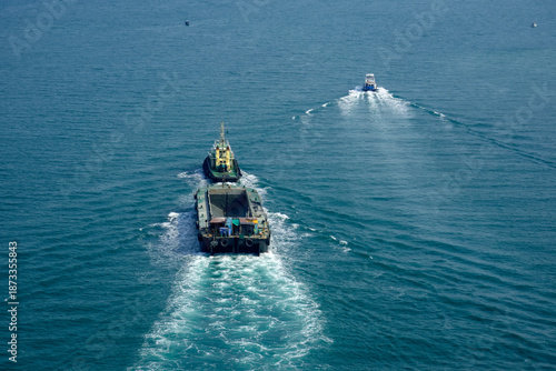 A tugboat pulls a cargo ship through calm waters close to the shore. The sky is clear, and the vessels create wakes as they move together. The scene shows activity on the water