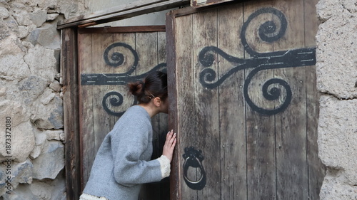 Fotografija A brunette girl in a gray knitted sweater with openwork sleeves looks into an old wooden door surrounded by stone walls