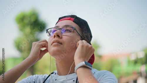 closeup portrait adjusting earbuds in park, reflective profile with glasses and backward cap, meditator centering before run, student calibrating audio for podcast, listener enjoying ambient nature