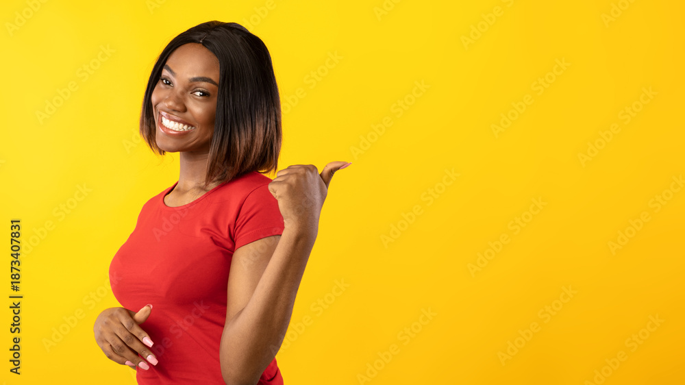 Obraz premium Look At This Offer. Cheerful Black Woman Pointing Thumb Aside At Free Space For Text Behind Her Back Standing Posing Over Yellow Background. Studio Shot, Panorama