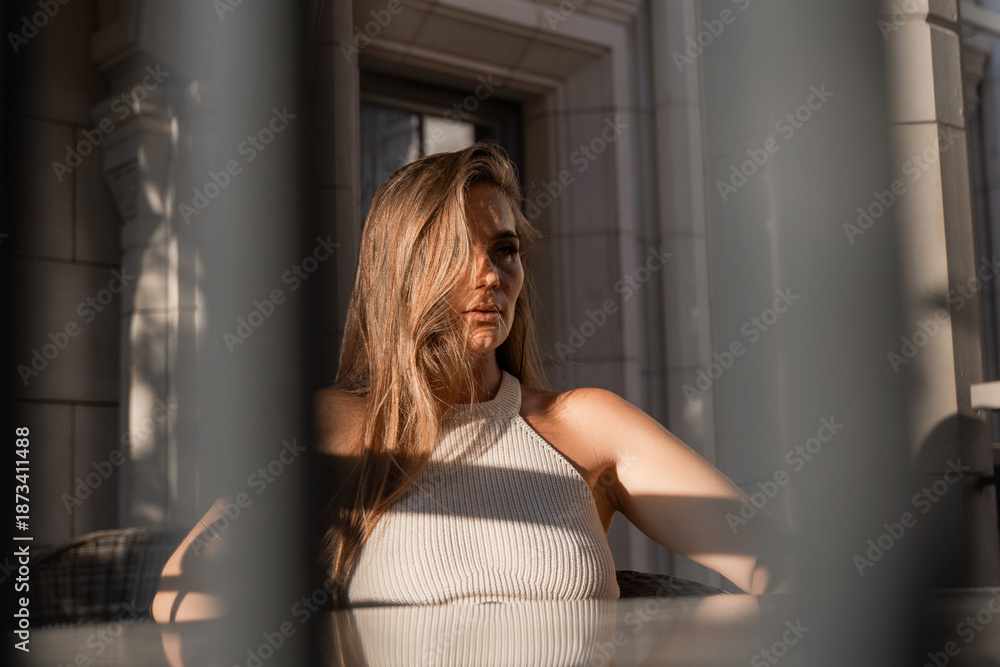 Fototapeta premium Woman, Sunlight, Window. Blonde woman sits at a table outdoors, bathed in warm sunlight streaming through a window.