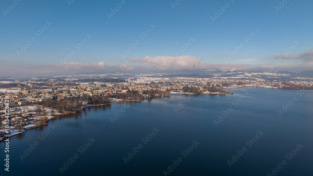 Fototapeta premium Aerial drone view along the shore of Hünenberg See shows snow-covered homes and residential blocks beside the deep blue Lake Zug under a clear, sunny winter sky.