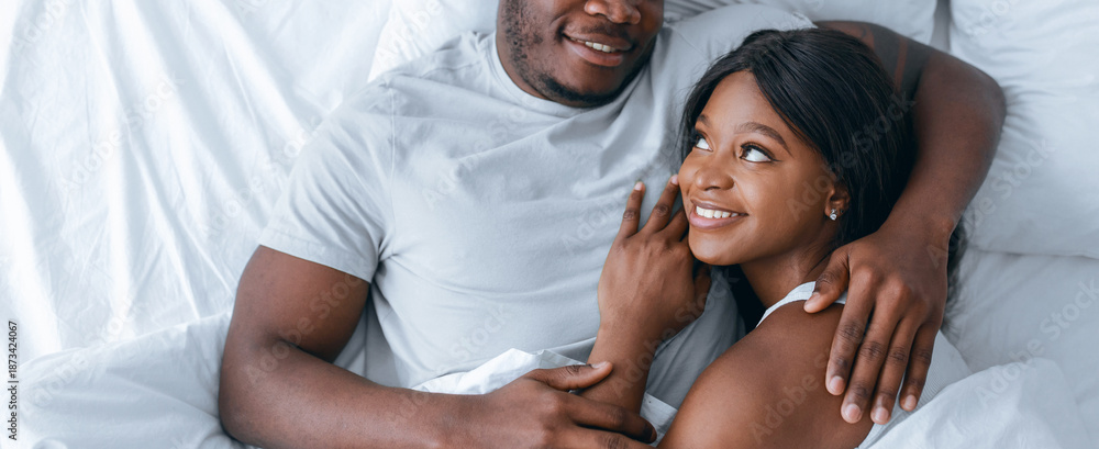 Fototapeta premium Two people lie together in bed, smiling and looking at each other. They appear happy and comfortable in their private space. Soft bedding surrounds them as they share a moment.