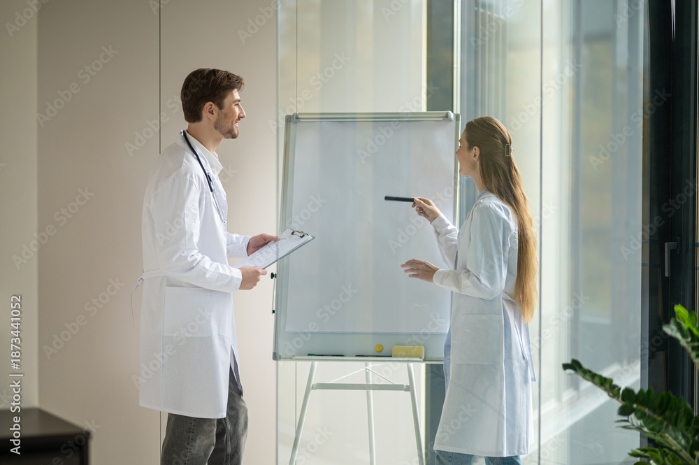Obraz premium Female doctor standing near the flipchart and presenting something to a new doctor