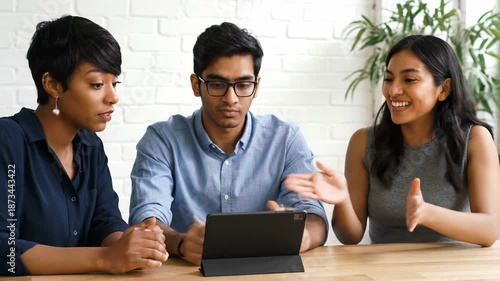 Diverse young professionals in smart-casual attire focus on a tablet at a light wood table in a bright indoor space with brick wall and plants, a cinematic video with shallow depth of field and