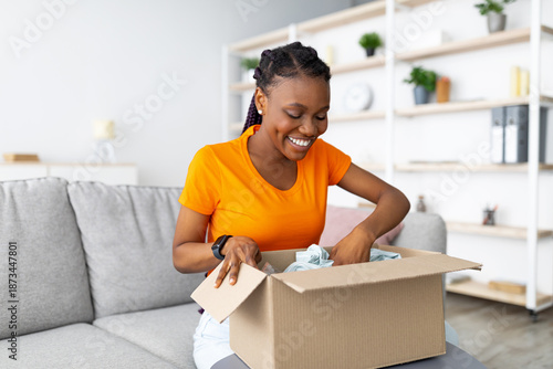 Online delivery service. Cheerful African American woman unpacking parcel box with purchase at home. Happy female customer satisfied with ordered goods, looking at new clothes, shopping on web