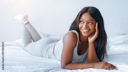 A woman is lying on her stomach on a bed, smiling and resting her head on her hand. The room is bright and has a soft atmosphere. She appears relaxed and comfortable.