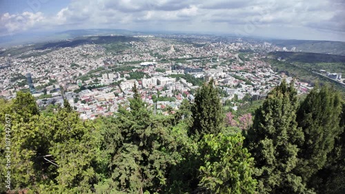 Lush greenery framing Tbilisi's cityscape reveals a blend of traditional and modern architecture, with the Kura River winding through the vibrant urban landscape under a sunny sky