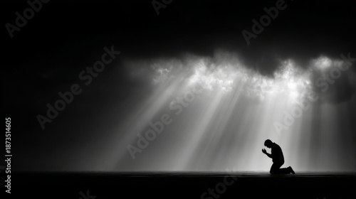 Black and white silhouette of man kneeling in prayer with dramatic light rays breaking through dark clouds creating spiritual atmosphere
