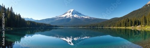 Wide view of calm lake reflecting snow-capped Mount Hood and dense evergreen forest under clear blue sky. Scenic mountain landscape with turquoise clear water and tree-lined shores.