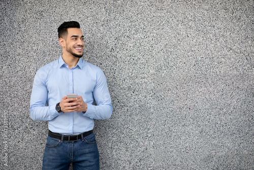 Фотография Handsome arab guy in formal wear standing over grey wall, holding cellphone and