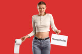 Young woman holding paper with word HEMORRHOIDS and roll on red background