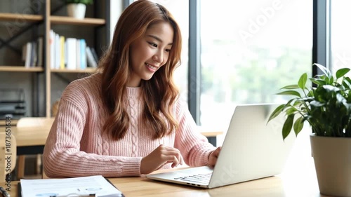 Asian woman working on laptop in bright, modern office, smiling and typing, conveying productivity and focus.