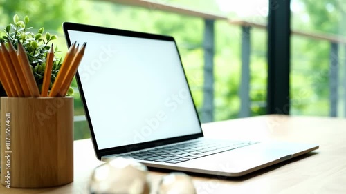 Open laptop with blank white screen on a wooden desk next to a pot of pencils and a green plant, with a blurred background of trees and a balcony railing.