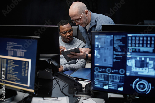 Caucasian man showing digital tablet to young Black man while sitting at workstation with multiple computer monitors, displaying technical data in dark office