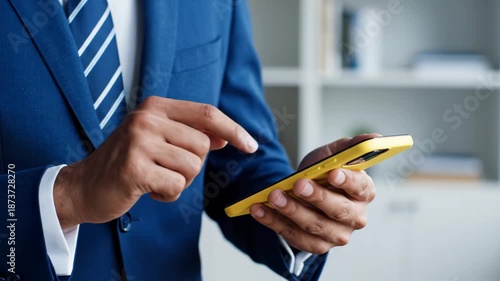 Close-up of a Professional Businessman in a Blue Suit Using a Smartphone with a Yellow Case in an Office, Browsing and Messaging on a Mobile App