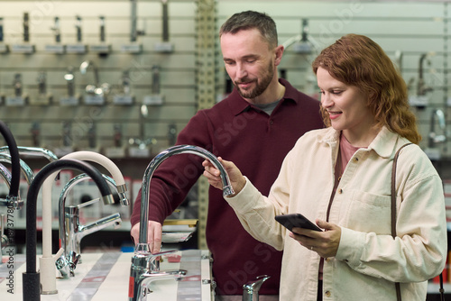 Caucasian man and Caucasian young adult woman examining kitchen faucets in store showroom, woman holding smartphone while both inspecting fixtures on display