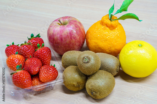 Assortment of fresh fruits on a wooden table. Strawberries in a plastic pack, red apple, kiwi fruits, Shiranui citrus and yellow orange. Healthy dessert ingredients.