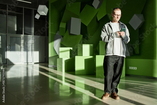 Wallpaper Mural Caucasian young adult man standing in modern office interior using smartphone, looking at device with focused expression, geometric green wall installation in background, sunlight streaming in Torontodigital.ca