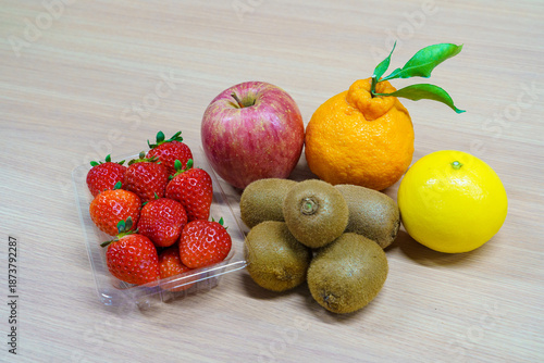 Assortment of fresh fruits on a wooden table. Strawberries in a plastic pack, red apple, kiwi fruits, Shiranui citrus and yellow orange. Healthy dessert ingredients.