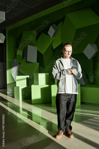 Young adult Caucasian man standing indoors using smartphone, looking down at device with neutral expression, geometric green abstract wall and cubes in background