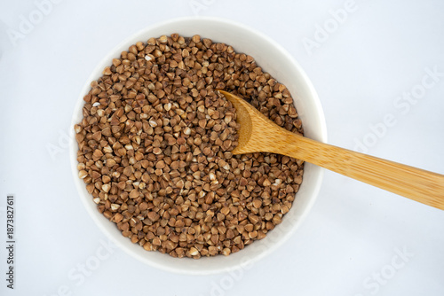 Top view of raw buckwheat groats in a white ceramic bowl with wooden spoon on a neutral background. Healthy food concept, clean minimal still life for nutrition and diet themes. 