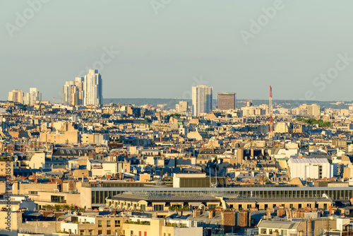 Expansive view over Paris rooftops with a mix of historic low-rise buildings and distant modern skyscrapers, bathed in warm evening sunlight under a clear sky.