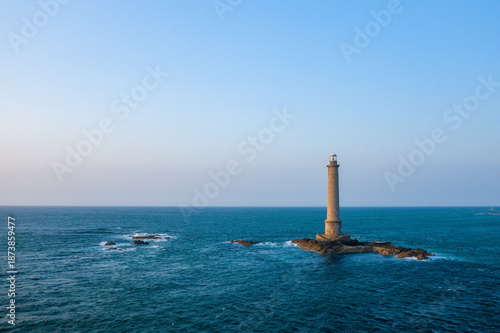 A tall lighthouse stands alone on a small rocky outcrop surrounded by deep blue sea at Cap de la Hague. The scene is illuminated by clear daylight, with gentle waves and a vast open sky creating a