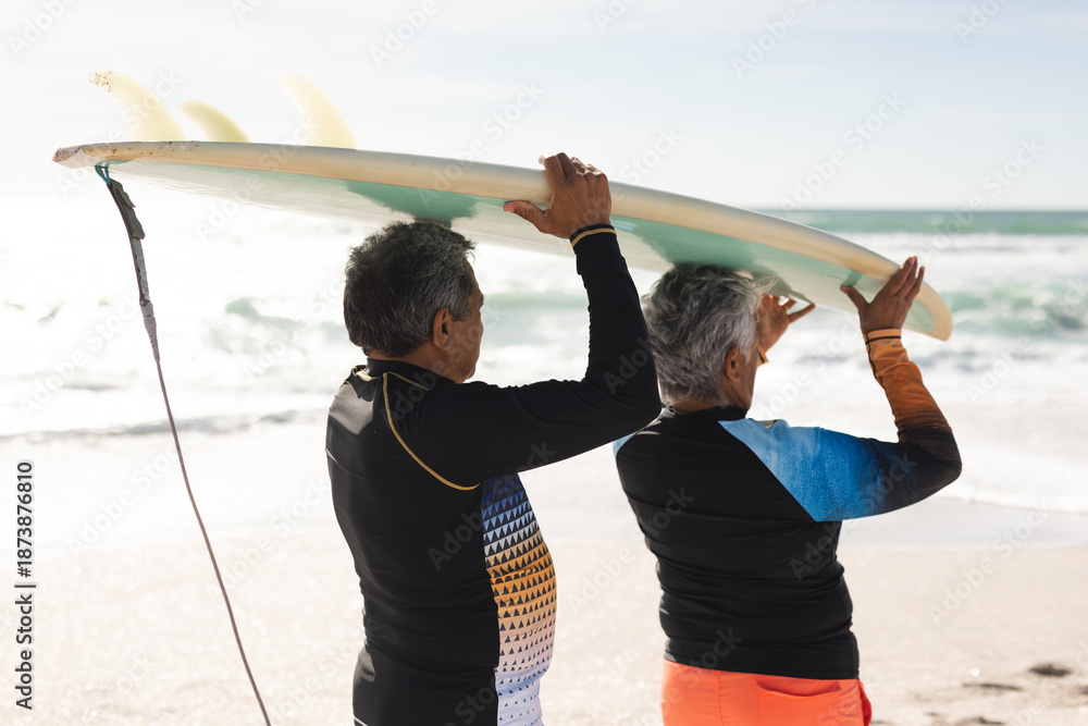 Fototapeta premium Multiracial senior couple walking together carrying surfboard over heads on shore at sunny beach