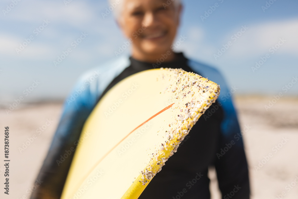 Fototapeta premium Surfboard with sand held by smiling senior woman at beach during sunny day
