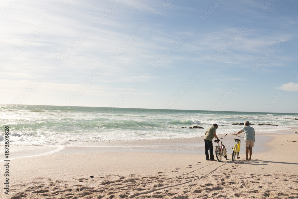 Naklejka premium Full length of multiracial senior couple walking with bicycles on shore at beach during sunny day
