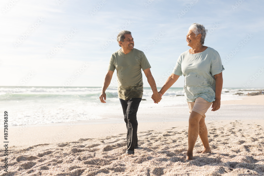 Fototapeta premium Happy senior multiracial couple holding hands while walking on sand at sunny beach against sky