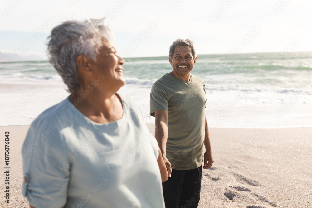 Fototapeta premium Smiling senior multiracial couple holding hands while enjoying sunny day on shore at beach
