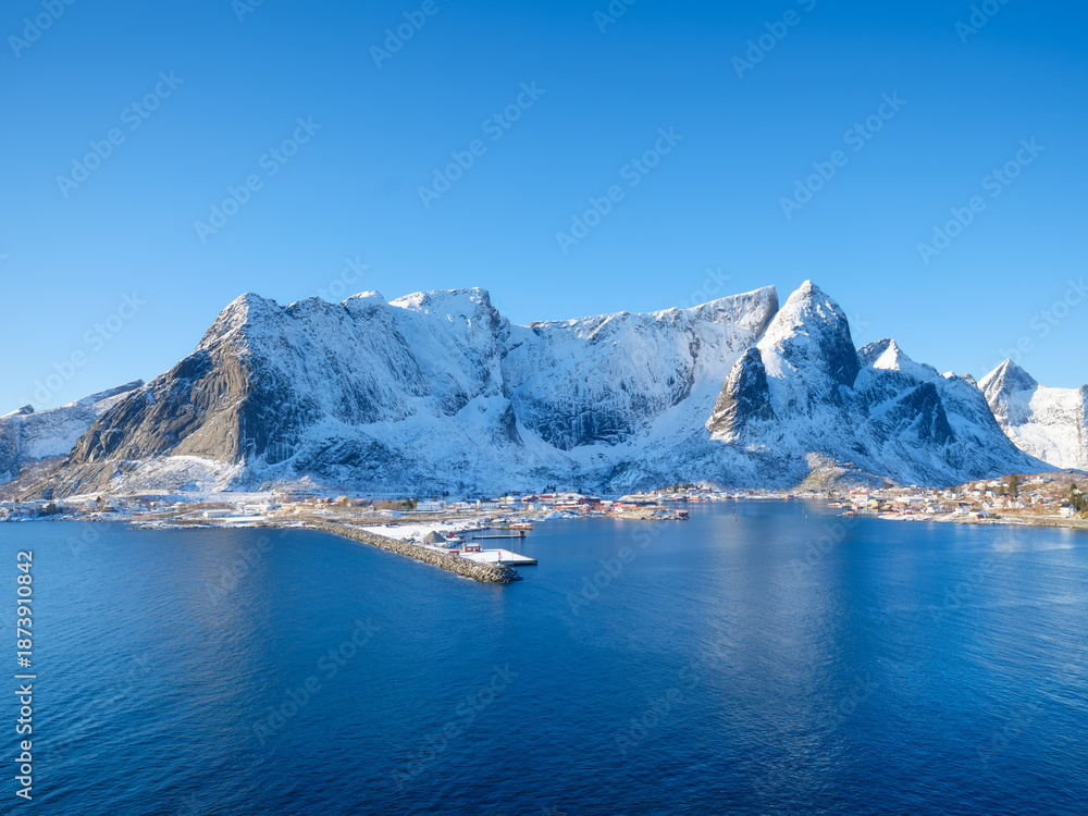 Obraz premium High arctic mountains and steep cliffs rising above deep fjord in winter. Majestic snowy peaks reflecting on calm water surface under bright sunny sky, Lofoten Islands, Norway.
