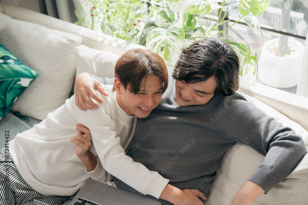 Obraz premium Relaxed Asian gay couple sitting on living room sofa and watching laptop together. A warm domestic lifestyle moment showing digital connection, comfort, and modern home entertainment for two men