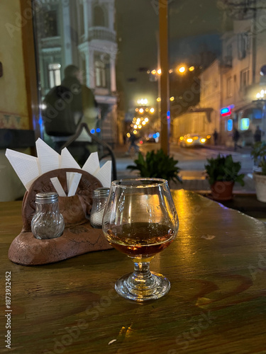 Glass of amber liquor rests on a wooden cafe table, glowing softly against a rainy city nigh