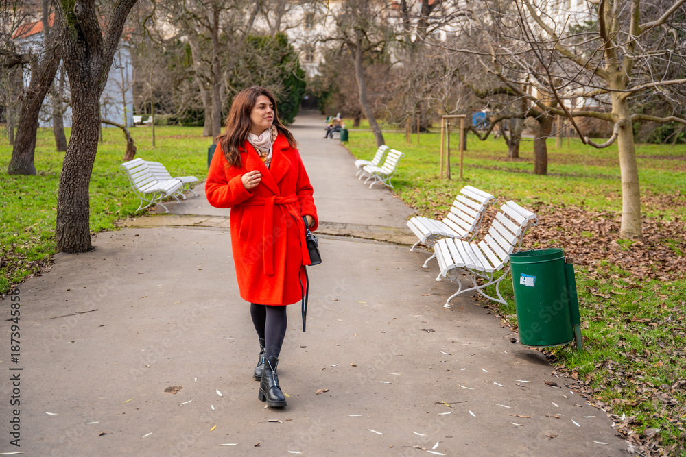 Fototapeta premium Woman walking in park in Prague during winter without snow wearing red coat. Calm urban walking, everyday lifestyle and seasonal city routine in real life European environment