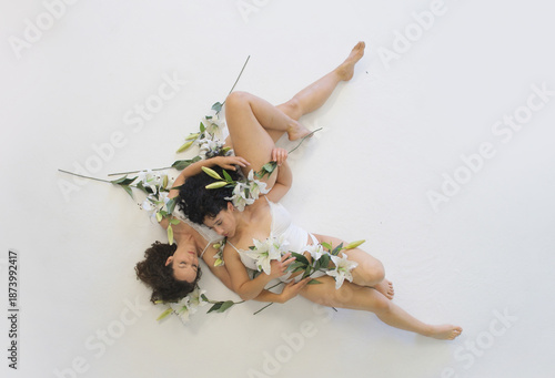 Two sexy women, curly hair, lie intertwined on a white floor, surrounded by lilies. The image evokes tenderness, intimacy, and purity, with a serene, peaceful atmosphere between them