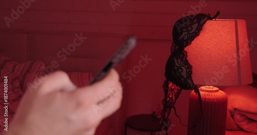 A person uses a smartphone in a red-lit bedroom while lingerie hangs on a lamp, suggesting private messaging, online flirting, and discreet romantic communication.
