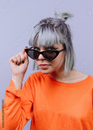 Young woman with light hair wearing black sunglasses and an orange top against a purple background