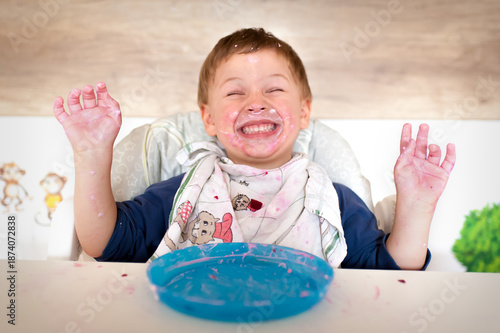 A child sits in a high chair covered in food while smiling widely. Colorful food is splattered on the table and his hands. The scene shows joy and delight during meal time.