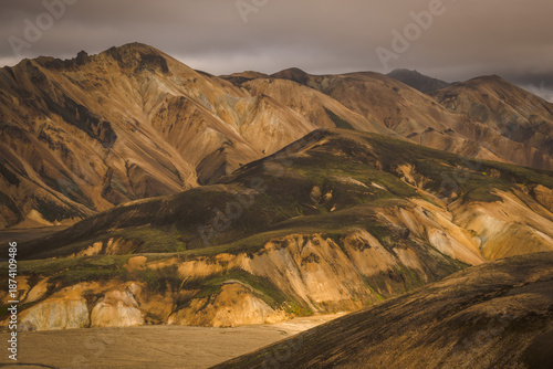 View of rugged, golden mountains rise under a brooding sky, their slopes patched with green, creating a stark contrast of color and texture, Landmannalaugar, Iceland.