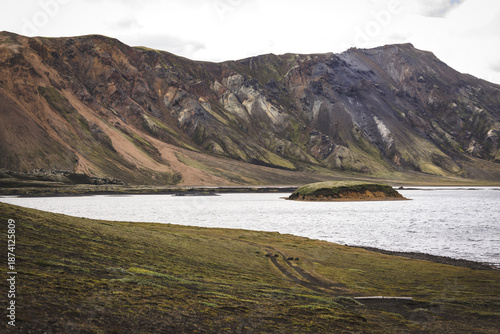 View of rugged, multicolored mountains rise majestically above a serene lake reflecting the cloudy sky, with a small island in the distance, Landmannalaugar, The Central Highland, Iceland.